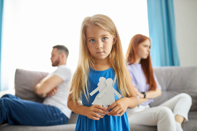 Little girl holding 2 paper cut outs of people with her parents sitting on the couch with their backs facing each other