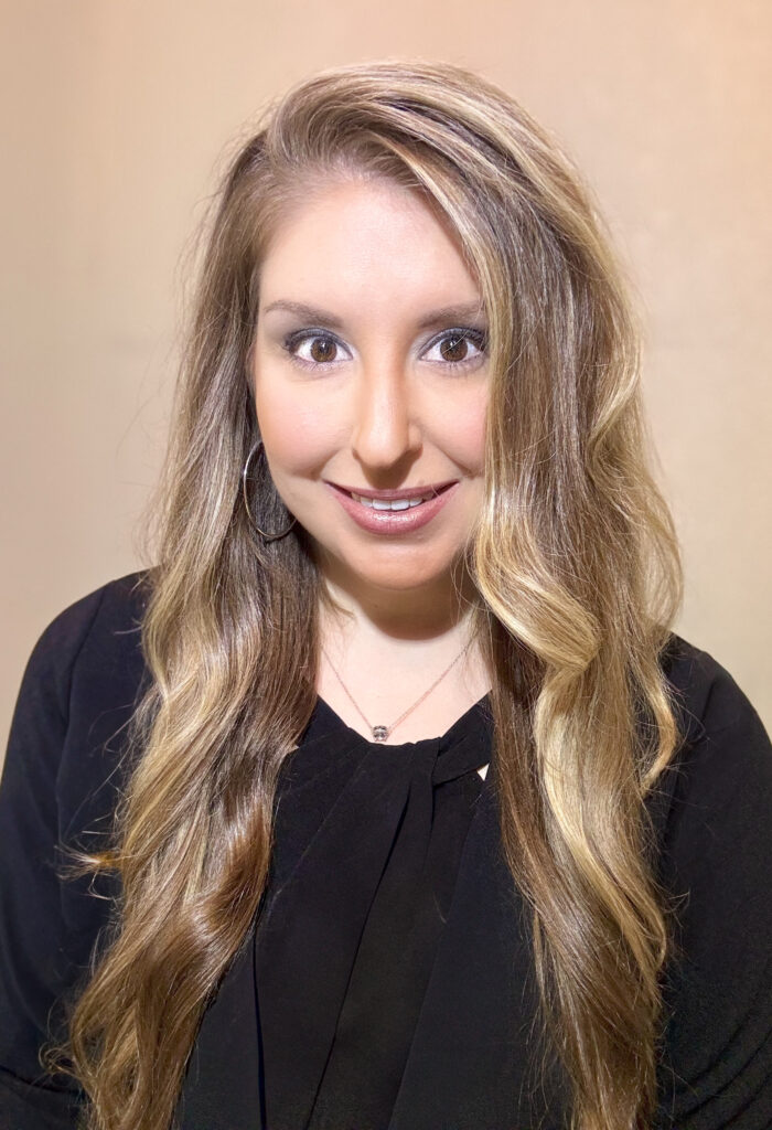 A woman with long, wavy blond hair smiles at the camera, wearing a black top.