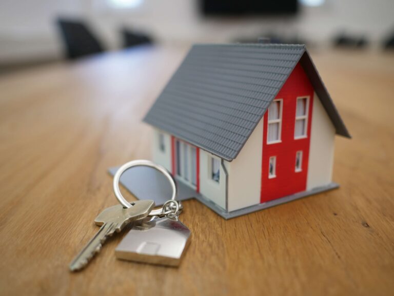 Little red and white house sitting on a wood table with a key attached to a house keychain next to it