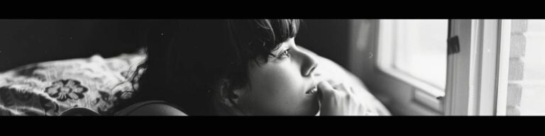 Black and white image of a person with short, dark hair resting their chin on their hand, showing signs of contemplation as they gaze out of a window, possibly reflecting on divorce or thoughts about their spouse.