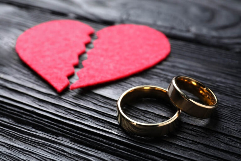 A red broken heart sitting next to two wedding bands on a dark wooden table