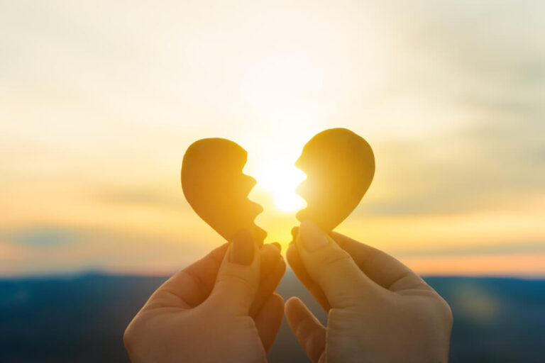 A heart cut into 2 pieces and a woman is holding the heart in front of a sunset and the woman has one piece in one hand and the other piece in the other hand