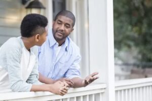 Two people are talking on a porch. One, wearing a light blue shirt, is gesturing with his hand, while the other, in a gray and white shirt, listens attentively about children's preferences in custody under Texas family law. Learn at what age can a child decide which parent to live with in Texas.