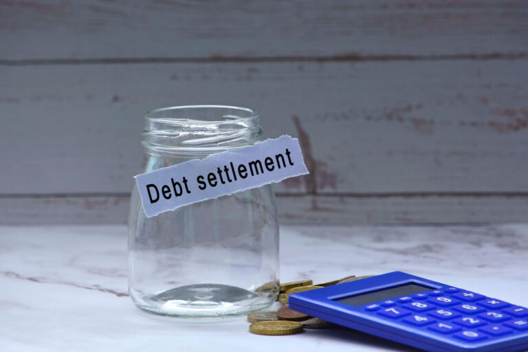 An empty jar with a paper that says debt settlement stuck to the jar and next to the card there are some coins on the table with a calculator on top of them