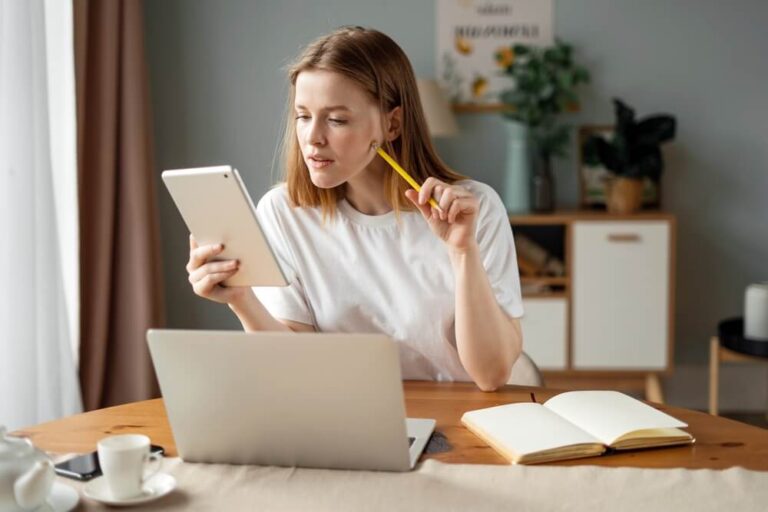 Women sitting at a table with her computer open and a notebook open next to the computer in one hand she has a tablet she is looking at and in the other hand she has a pencil