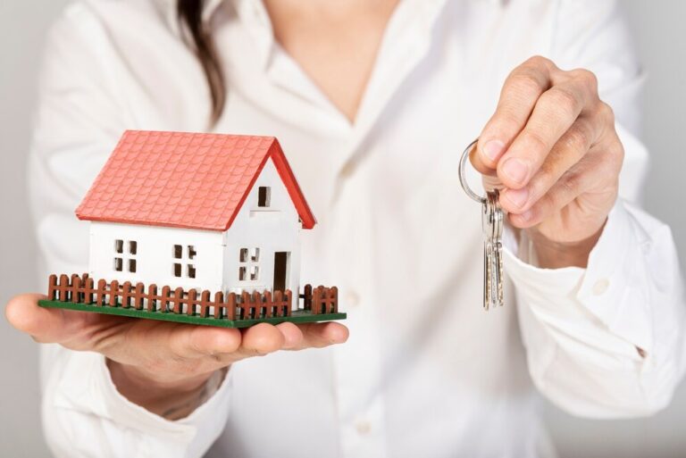 Person holding a small model house in one hand and a set of keys in the other, symbolizing real estate or home ownership, leaving some to ponder: does a will override a trust?