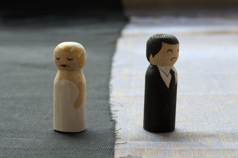 Two wooden figurines, one representing a woman in a white dress and the other a man in a black suit, stand apart on a surface divided by a seam. Their separation emphasizes dealing with an unreasonable ex post-divorce, highlighting the emotional distance.