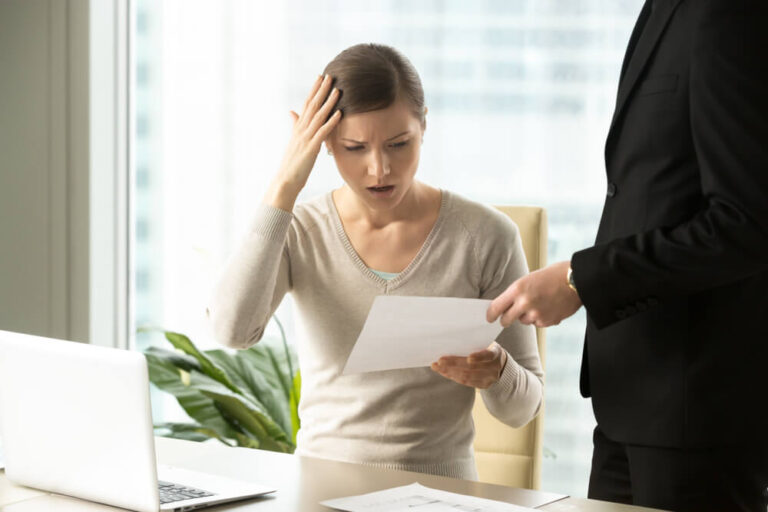 A woman sitting at a desk with a confused and shocked look on her face while being handed a paper