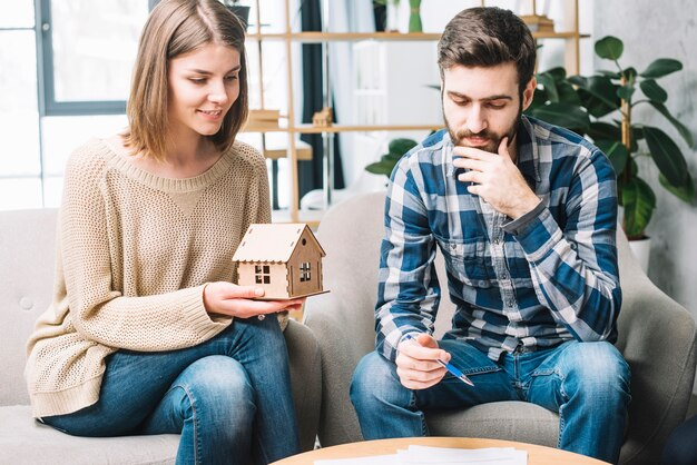 A woman holds a small house model while sitting beside a man  with various papers spread on the table in front of them.