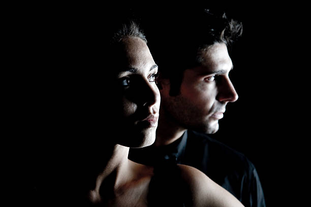 Two people are positioned back to back, looking in opposite directions, against a dark background with dramatic lighting highlighting their profiles.