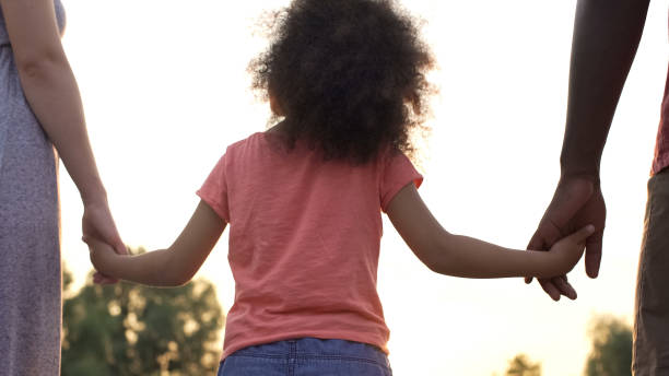 A child with curly hair, wearing a pink shirt, holds hands with two adults. Amid the beautiful Texas sunrise, these parents find joy together while pondering if they can agree to no child support in Texas.