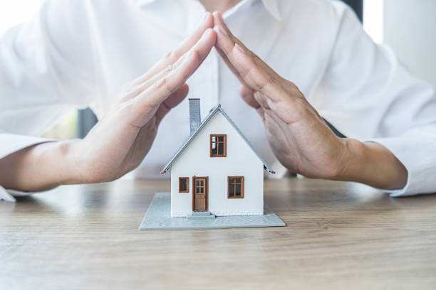 A person places their hands protectively over a small model house on a wooden table, symbolizing home security or insurance, akin to learning how to put a house in trust with a mortgage.