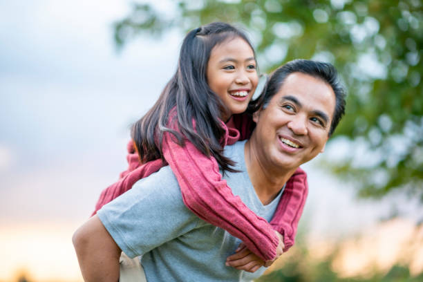 A Filipino father gives his elementary school aged daughter a piggy back ride, while they enjoy a walk outside. They are smiling and enjoying each others company and his daughter is looking down at him.