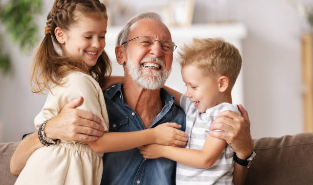 An older man with glasses and a white beard embraces two young children, a girl and a boy, while sitting on a couch. All three are smiling and appear to be happy, reminiscent of the cherished moments that grandparents rights in Texas aim to protect.