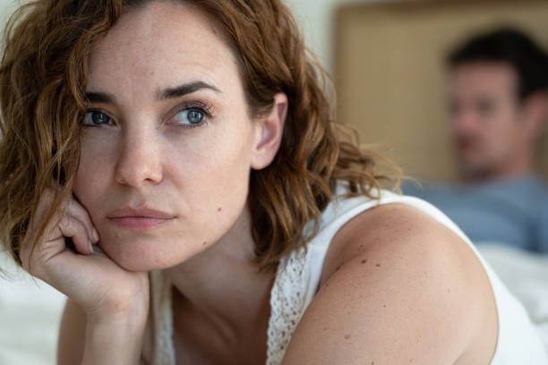 Sad woman sitting on the bed's edge while her partner lying on the bed and using a smartphone in the background