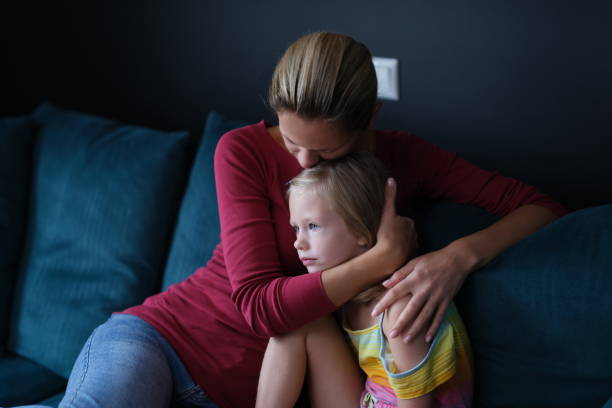 An adult in a red shirt hugs a young child in a multicolored dress on a blue couch against a dark background, both appearing deep in thought.