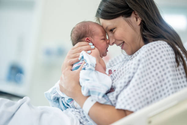 A new Mother sits up in her hospital bed shortly after delivery as she holds her newborn out in front of her and studies his features. She is wearing a hospital gown and is laying in her hospital bed with the inafnt.