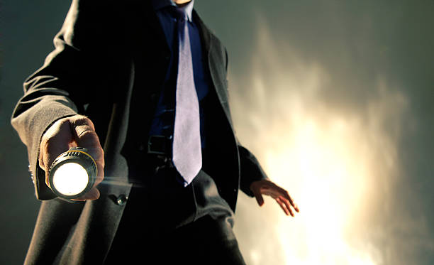 A close up of the torso of a Caucasian man dressed in a dark business suit, shirt and tie holding a battery operated torch pointed towards the camera, with his arm out in a dramatic pose. The man is standing against dark background with a brightly lit abstract pattern.