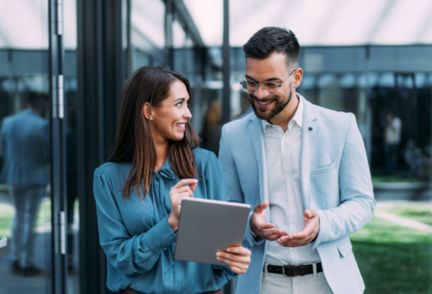 Two people in business attire stand outside a building, smiling as they review successful commercial real estate restructuring plans on a tablet.