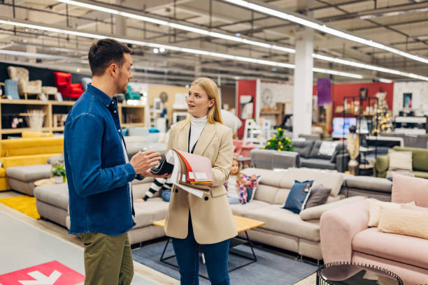 A carefree saleswoman in a wholesale furniture store shows samples of colors and materials to a smiling male customer, while his family sits on a sofa in the background