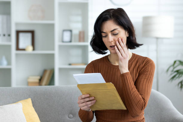 Close-up photo of worried and shocked young woman sitting on sofa at home and reading received letter and message, holding hand to head in shock.