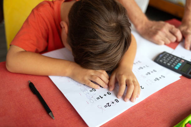 A six-year-old boy, depressed and frustrated, has his head down on his math homework sheet, clearly unwilling to continue. His father stands next to him, his hands on the table, supporting him and waiting for his son to be ready to continue.