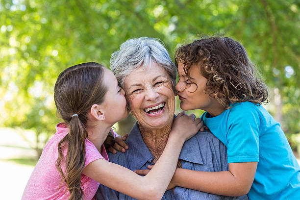 An elderly woman, exercising her grandparents' rights in Texas, is being kissed on the cheeks by two children, one on each side, while smiling. They are outdoors with green trees in the background.