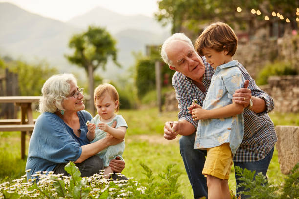 An older couple with gray hair spends time outdoors with two young children, one seated on the ground and the other standing, near a garden with green plants and flowers, embracing their grandparents' rights in Texas.