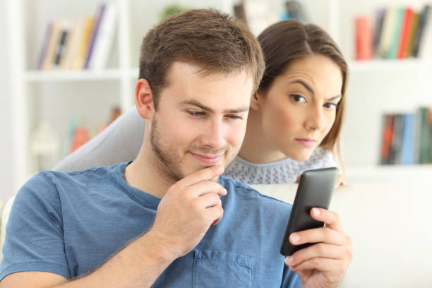 A man is looking at his phone and smiling while a woman, appearing suspicious or curious, looks over his shoulder. They are indoors with shelves and books in the background. The scene makes one wonder if virtual infidelity might be a ground for divorce.