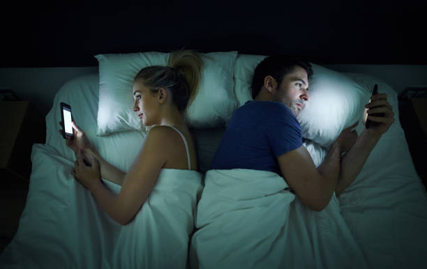 Shot of a young couple using their cellphones in bed at night back to back