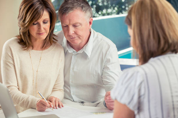 Senior Couple Meeting with Financial Advisor. They are listening to financial advisor. She is speaking about retirement, insurance and investment options. The mature woman is signing a document. Could be a will or a mortgage. The couple look worried