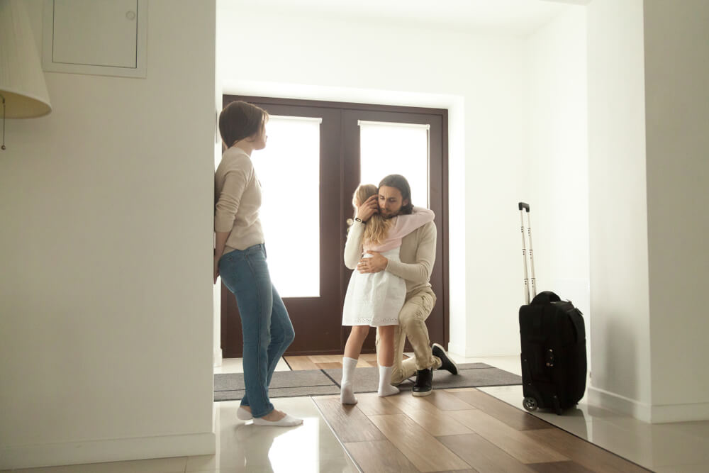 Father kneeling down to hug his daughter and mother is resting against the wall, there is a suitcase next to the father 