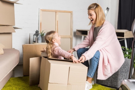 A woman kneels and smiles while interacting with a child sitting in an open cardboard box, surrounded by more boxes and furniture in a room, as if imagining the whimsical possibilities of their new home after sorting out child custody in Texas.