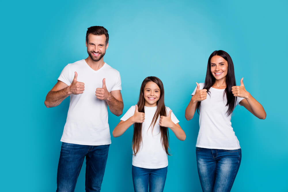 Father, daughter and mother standing side by side with their thumbs up against a blue background 