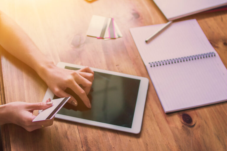 Person uses a tablet and holds a credit card next to a notebook and pen on a wooden desk.