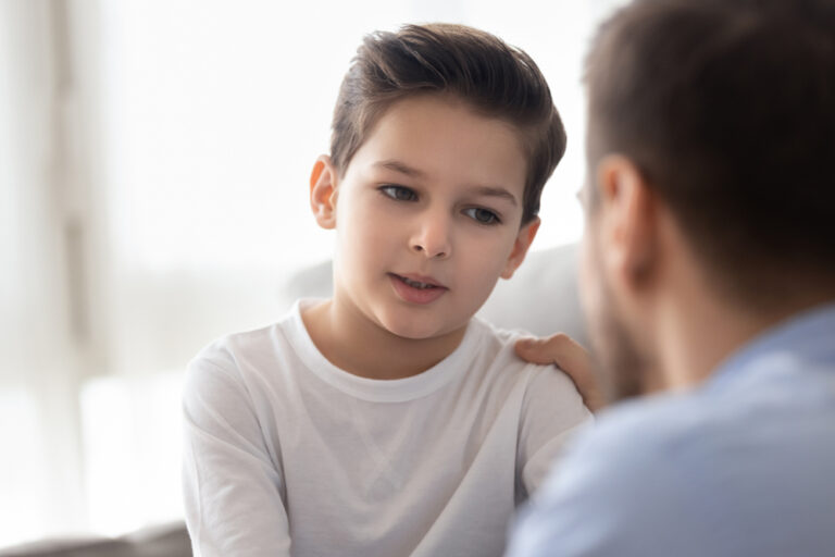 Back view of dad sit on couch with preschooler son talking sharing thoughts or problems, loving father speak have conversation with little boy child, support him showing care and understanding