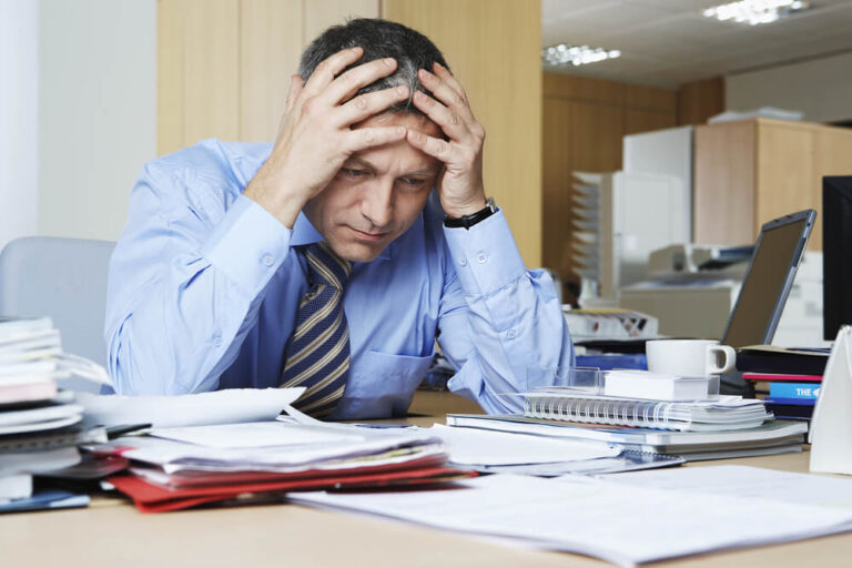 A man sitting at a table with many different stacks of papers on the table and his elbows are on the table with his hands holding up his head