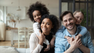 A smiling family of four, two adults and two children, pose together in a living room. One child is being held by each adult, and natural light fills the space.