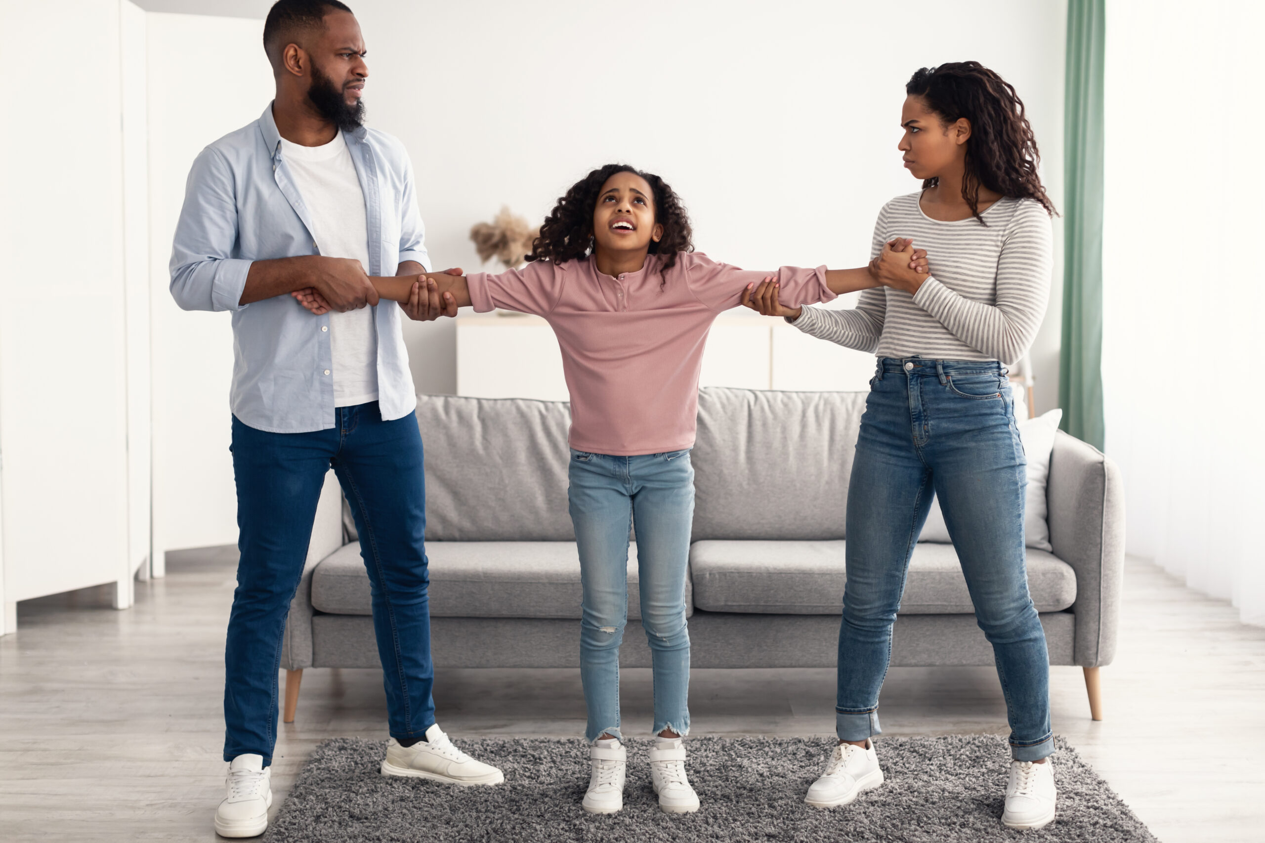 A young girl playfully stretches her arms out between two adults, who each hold one of her hands in a modern living room.