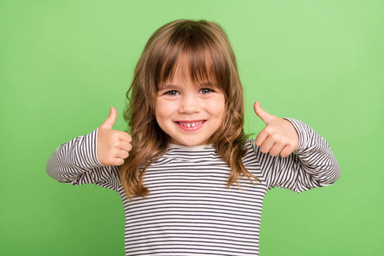 A smiling little girl standing against a green background with her thumbs up