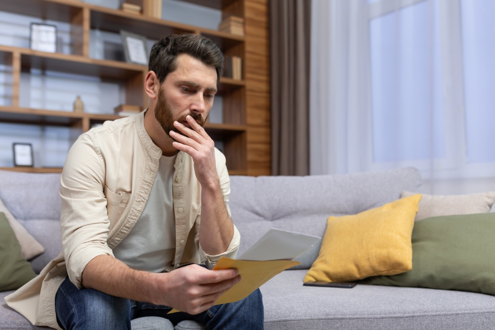 A man sitting on a couch reviewig legal documents wiht a stressful look on his face.