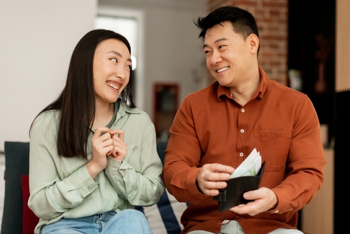 Two people sit on a couch, smiling at each other. The person on the right holds an open wallet with visible cash, perhaps pondering over matters like who pays attorney fees in divorce cases in Texas.