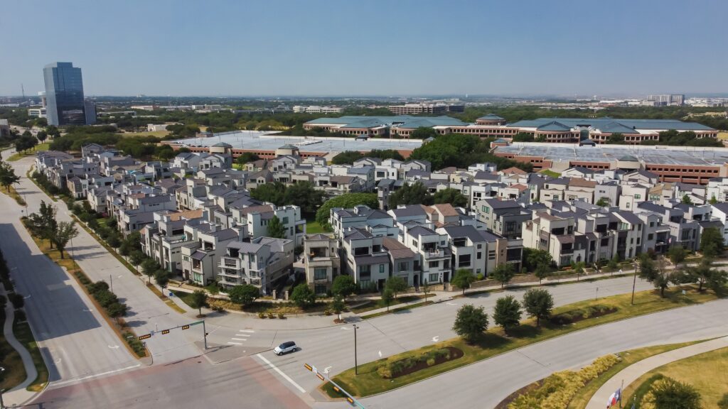 Aerial view of a Plano, Texas residential neighborhood with modern townhouses, a few office buildings in the distance, wide streets, and greenery around.