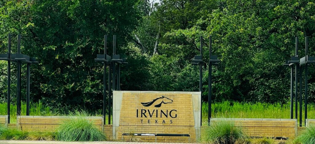 A brick sign surrounded by greenery reads "Irving Texas" with a stylized horse graphic above the text. Four decorative black posts flank the sign.