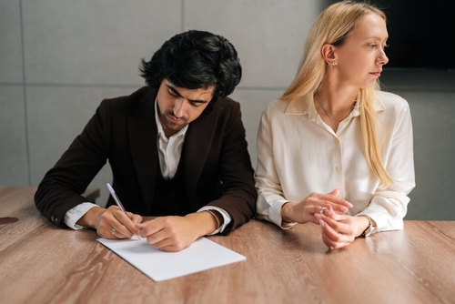 In an office setting, a man writes on a piece of paper while a woman sits beside him, looking away. The wooden table between them is scattered with documents detailing how to get divorce papers in Texas.