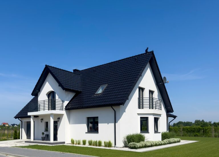 A modern white house with a black roof, featuring multiple windows and a small balcony. It is surrounded by a well-maintained lawn under a clear blue sky.