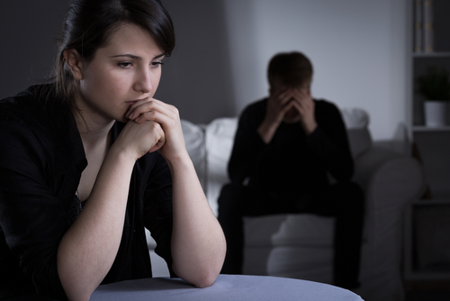 A woman sits in the foreground looking pensive, contemplating whether mediation is required for divorce in Texas, while a man in the background sits on a couch with his head in his hands.