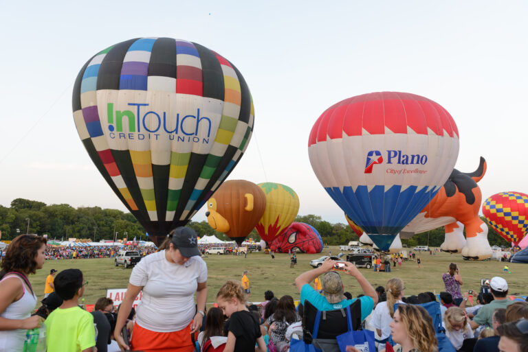 Hot air balloons, including some shaped like animals, inflate at a crowded festival with onlookers during sunset, near a banner advertising Plano divorce lawyers.