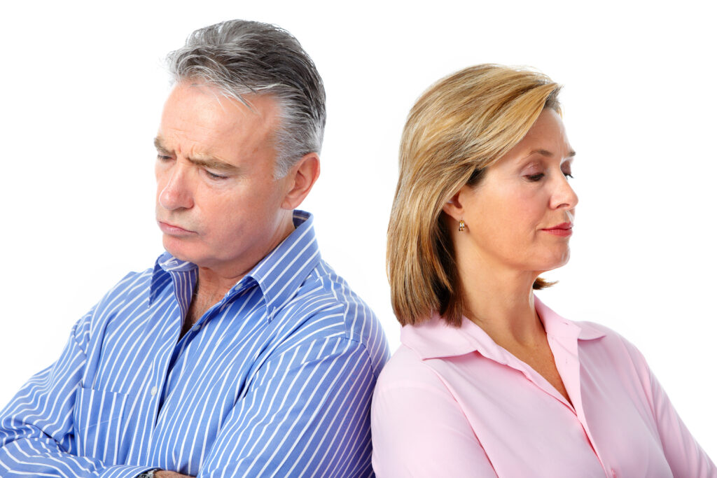 A man and a woman stand back-to-back with arms crossed, both looking away from each other, appearing upset. The man has grey hair and wears a blue striped shirt. The woman has blonde hair and wears a pink shirt.