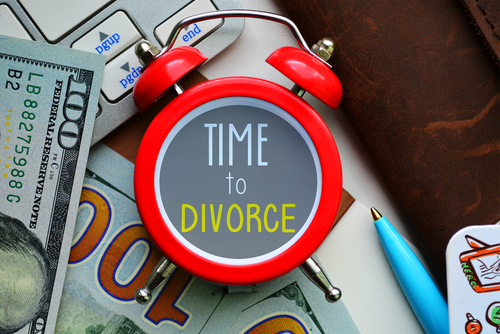 A red alarm clock with "TIME to DIVORCE" emblazoned on its face sits amid a hundred-dollar bill, a pen, and a leather item, silently questioning how long can a divorce be put on hold.